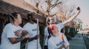 Photo of a group of 4 people in white volunteer t-shirts taking a selfie while holding brooms in a street 