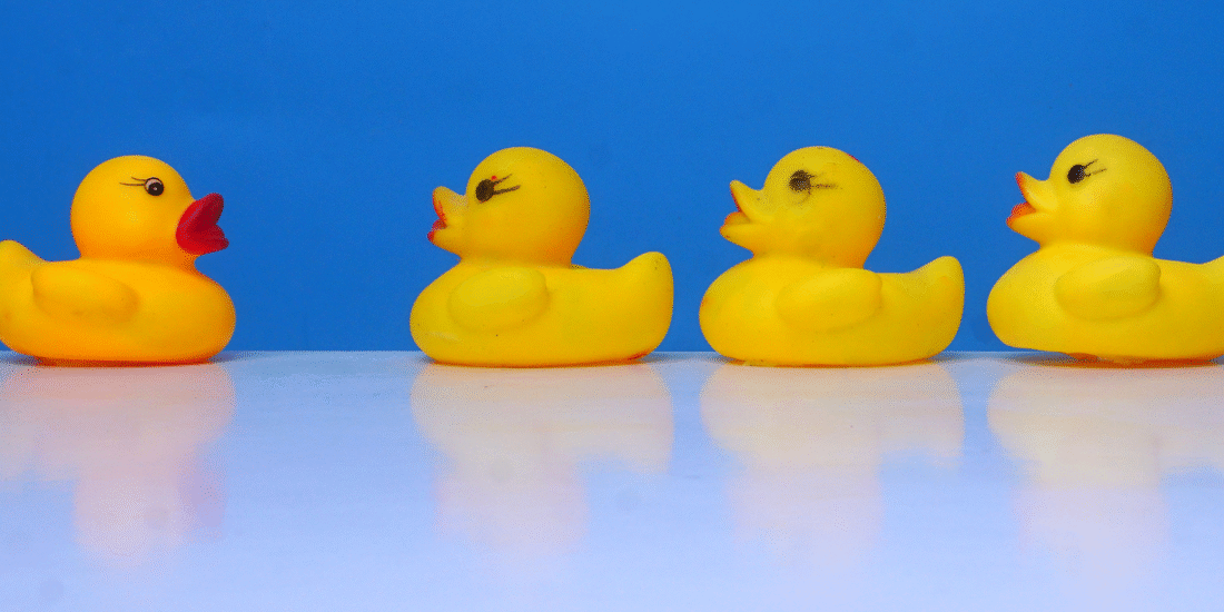 Yellow plastic duck facing a row of yellow ducks on a table with a bright blue background