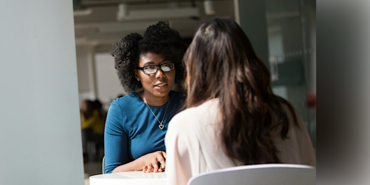 Woman in blue top and glasses talking across a table to woman in white top