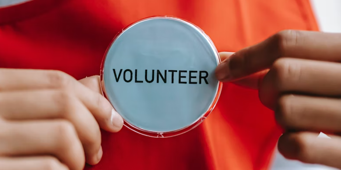 Close up photo of a white round badge with the word 'volunteer' in black typed capital letters, held by a pair of hands of someone wearing a bright orange t-shirt