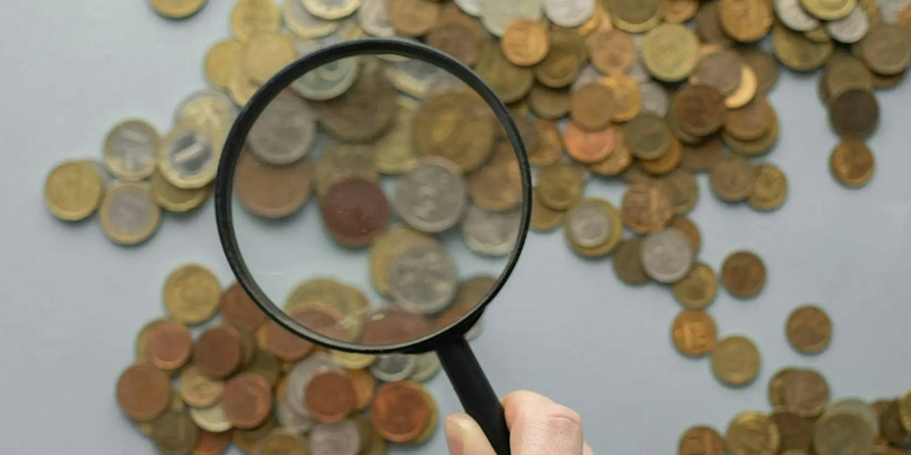 Hand holding a magnifying glass over scattered coins