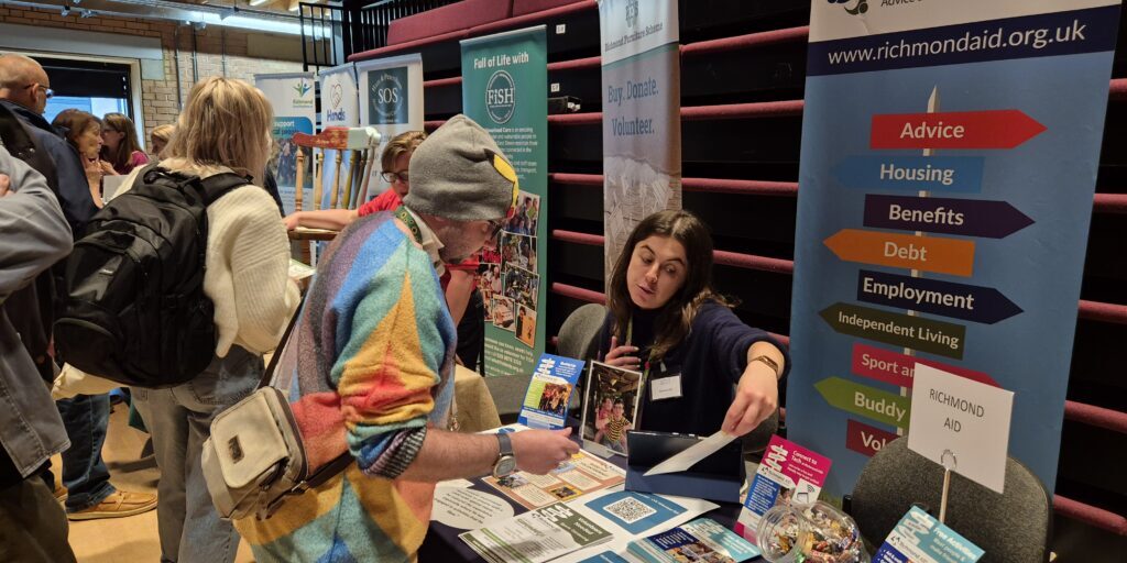 Visitor in hat and bright multicoloured jacket talking to charity stallholder at 2025 Richmond Volunteer Fair