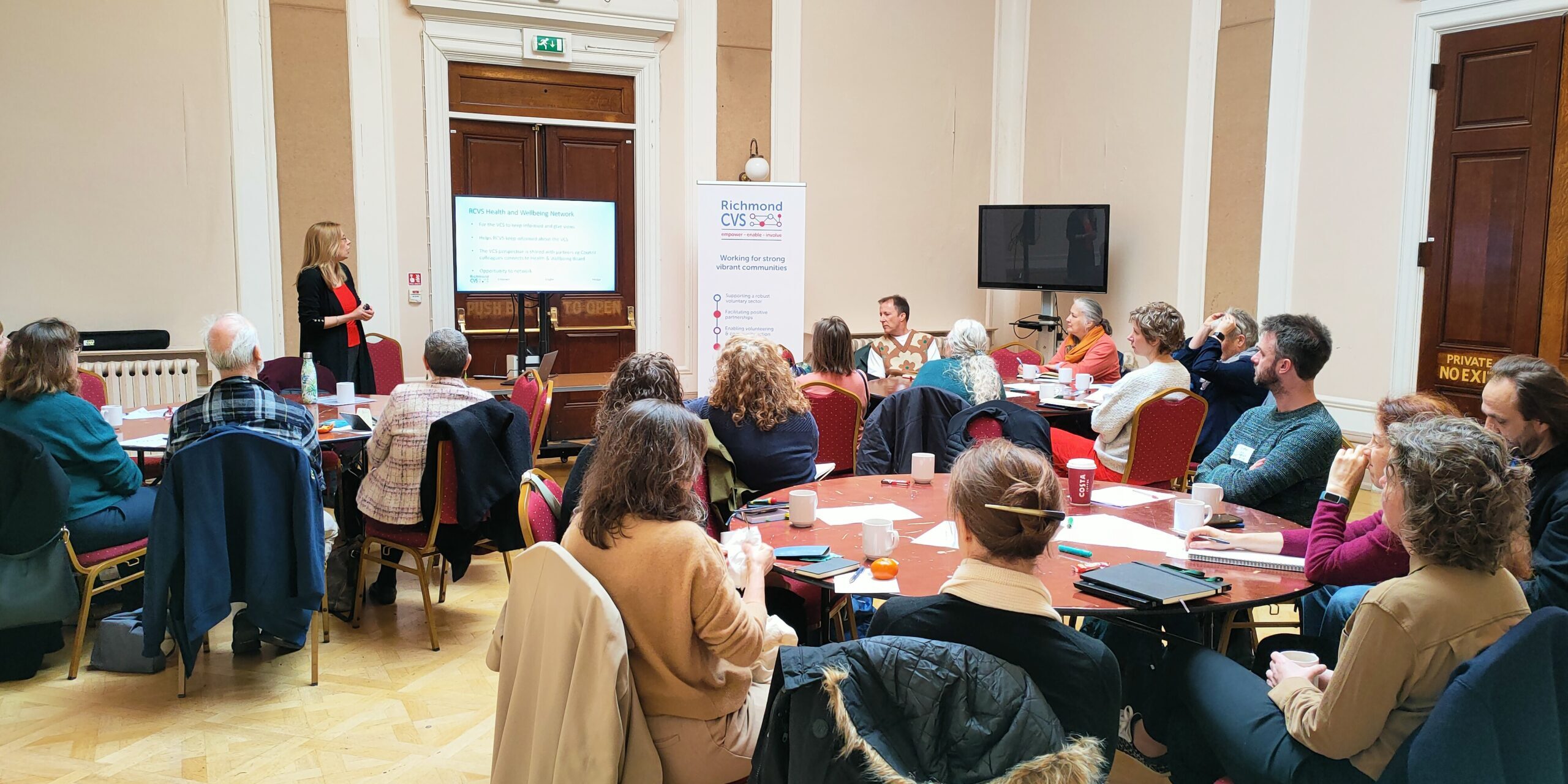 Photo of large room with seated attendees around tables and presentation screen and Richmond CVS banner at front of room