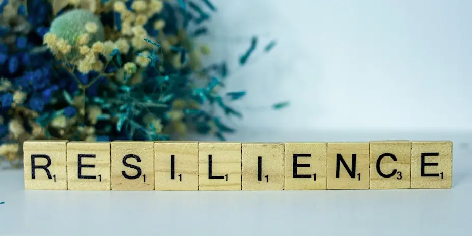 Wooden Scrabble pieces spelling RESILIENCE on white table with blurred bouquet of blue, green & yellow flowers background