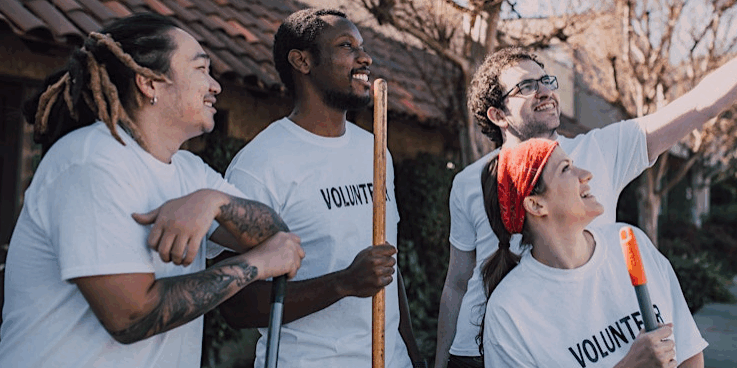 Mixed group of two young men and women in white t-shirts with black type 'Volunteer' holding brooms on a street and looking up to  take a selfie